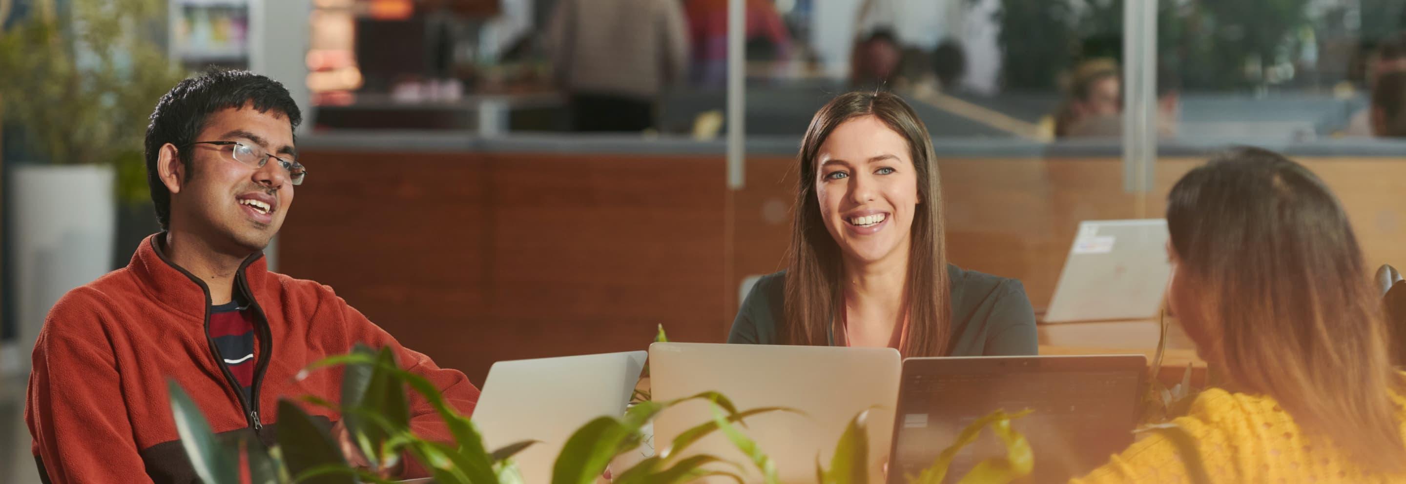 Three people with laptops smiling in conversation in a bright office with greenery in the foreground.
