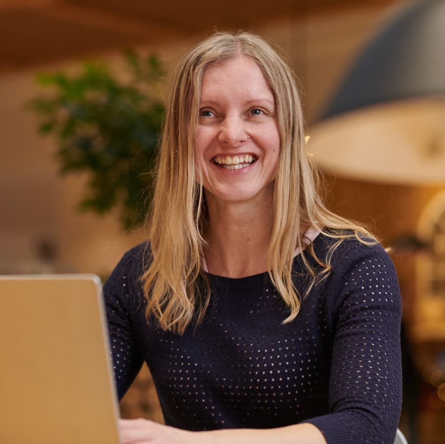 Smiling woman with long blonde hair using a laptop in a warm, modern office space with soft lighting.
