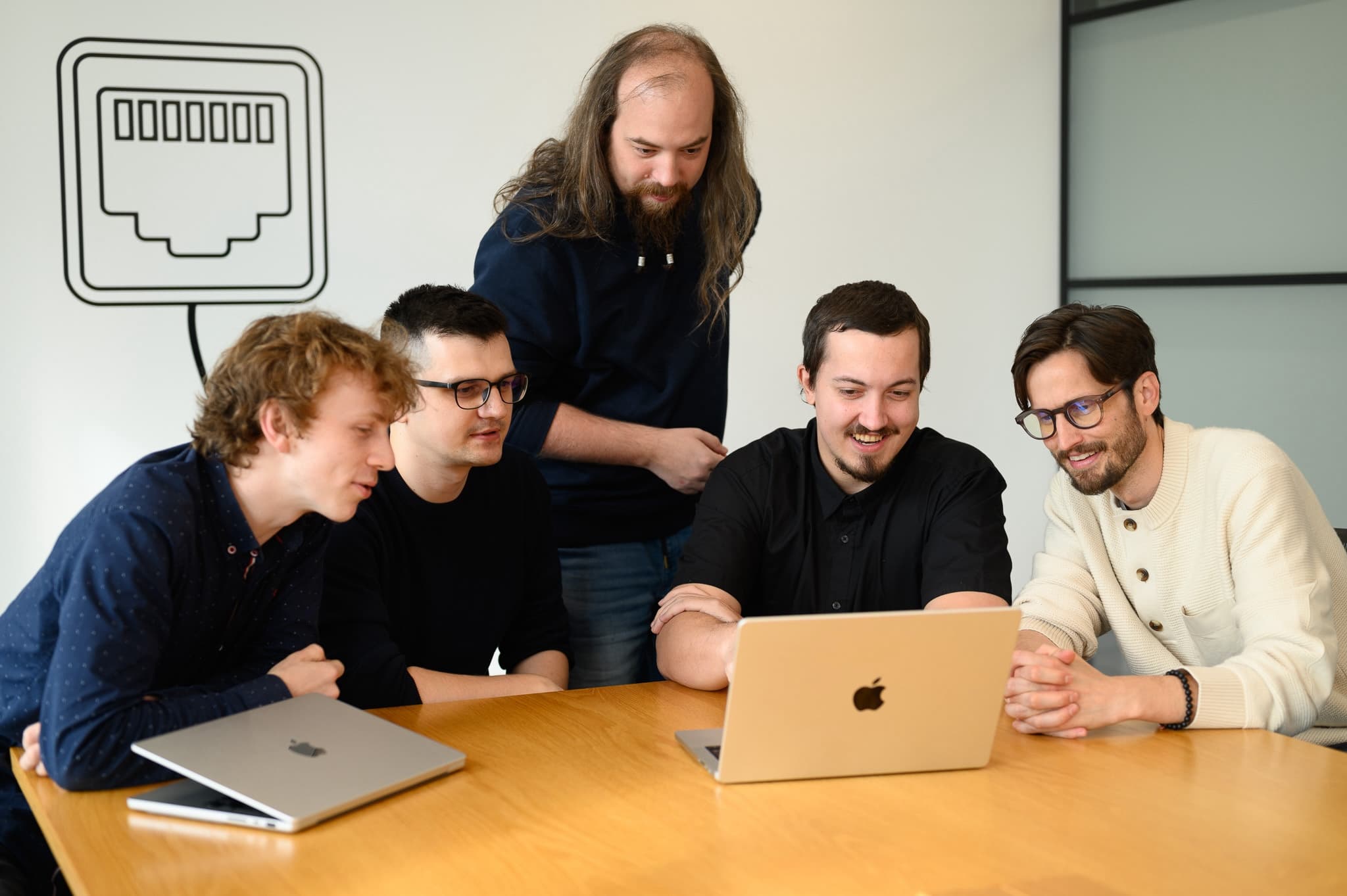 Five teammates gathered around a laptop, smiling as they collaborate in a creative workspace.