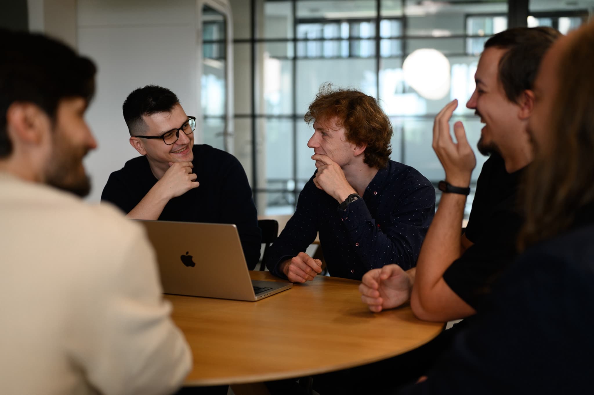 Group of colleagues laughing and chatting around a table in a bright modern office.