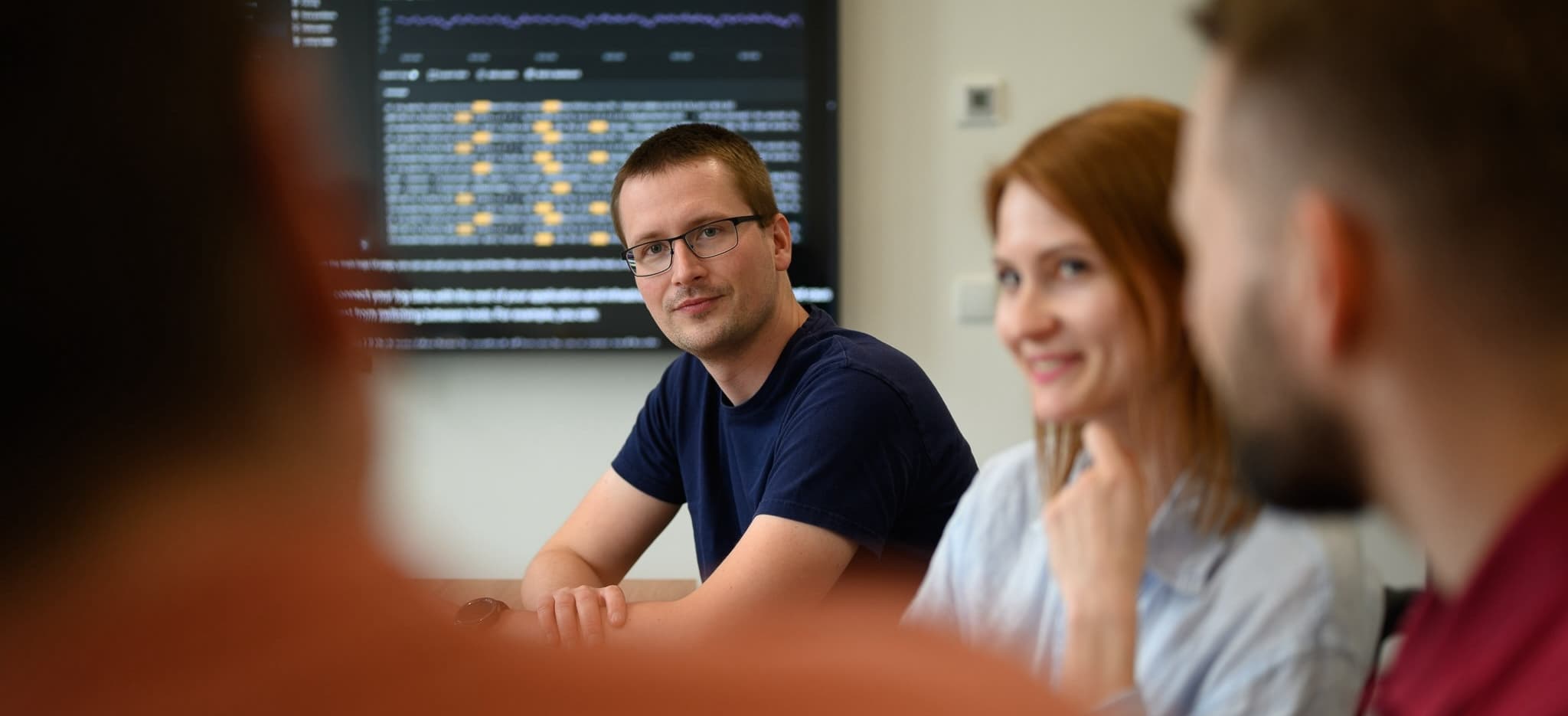 Man in glasses looks at the camera during a meeting with team members and a data screen.