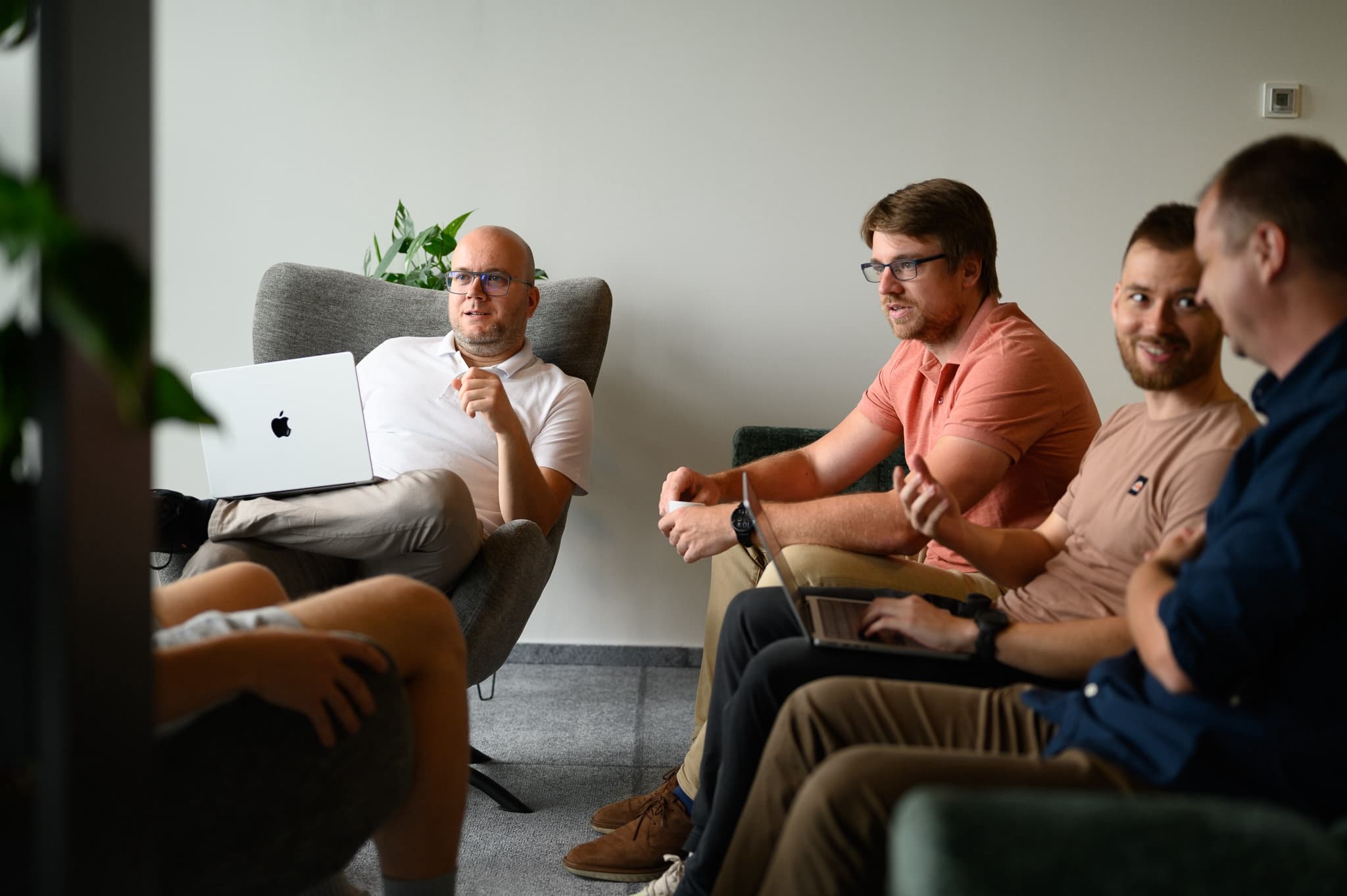 Four men in a casual meeting seated in lounge chairs, discussing something while working on laptops.
