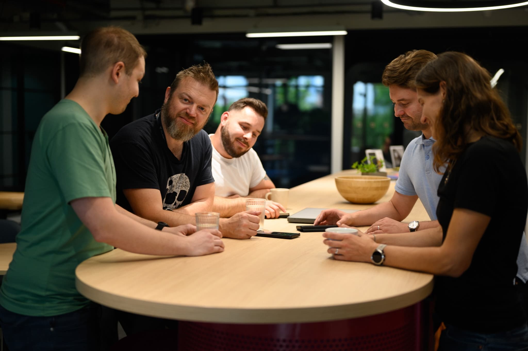 Five coworkers gathered around a round table with drinks and devices, chatting and smiling.