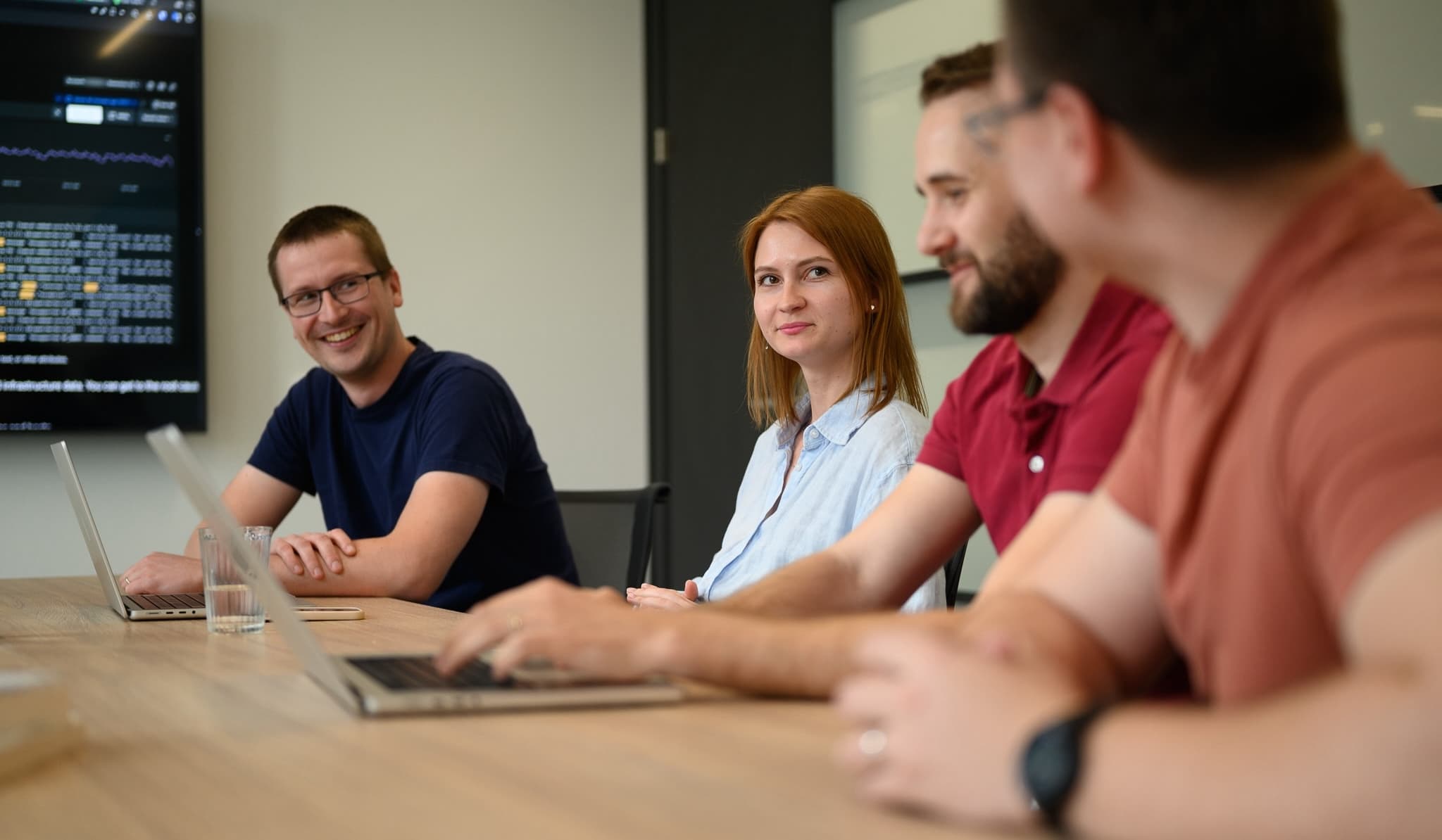Four colleagues sitting at a conference table with laptops, chatting and smiling in front of a data screen.