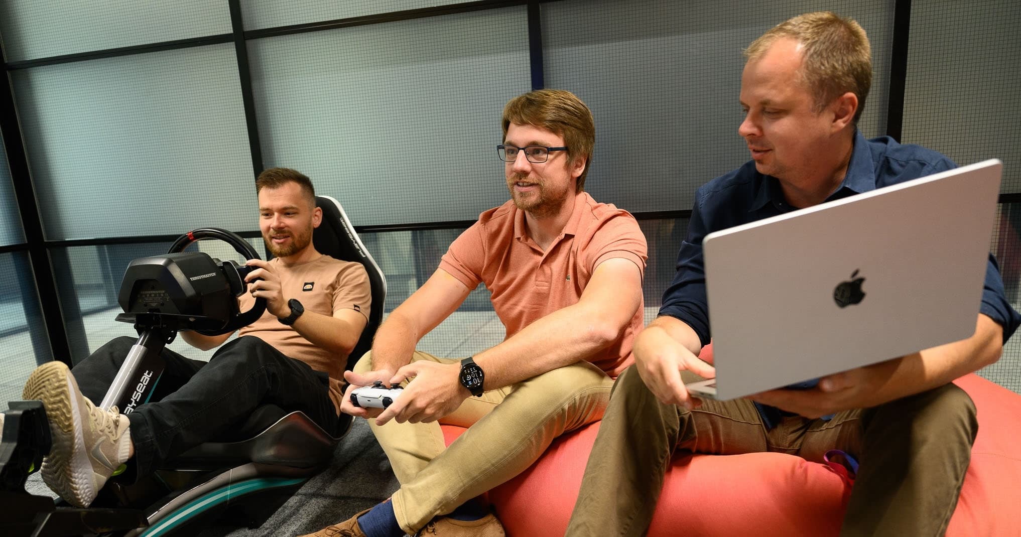 Three men casually playing video games on bean bags, one using a laptop, in a relaxed office space.