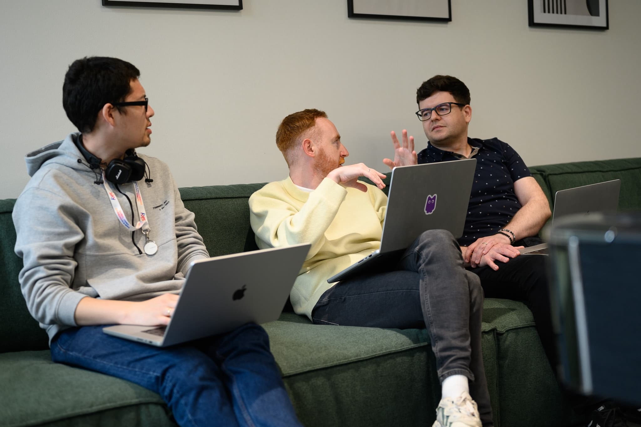 Three coworkers sitting on a green couch having a lively conversation, laptops open on their laps.