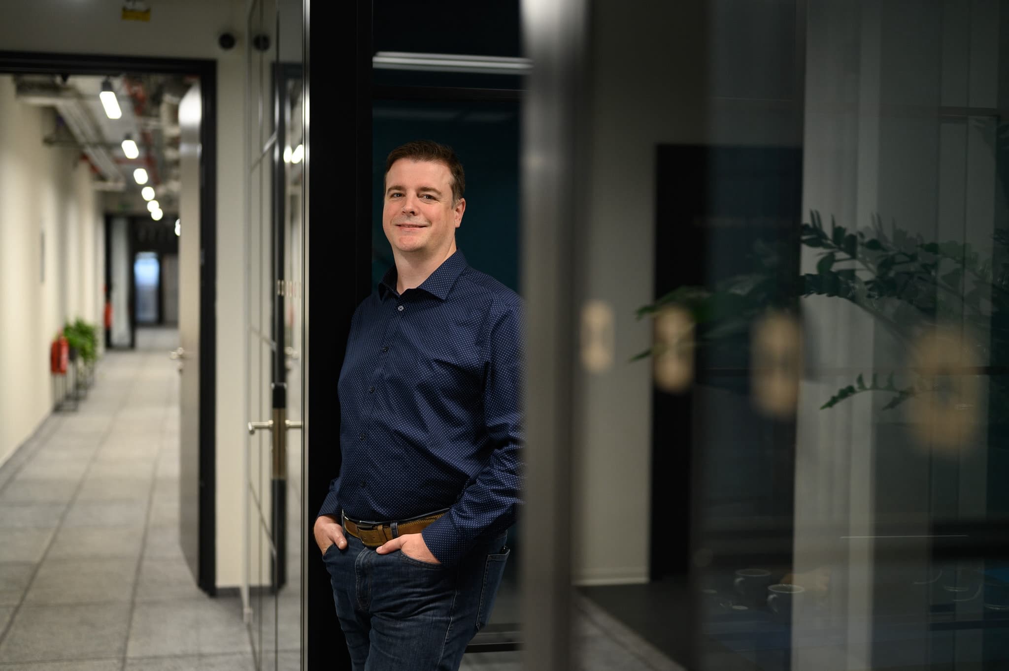 Smiling man in a blue shirt stands casually in a hallway, leaning against a door frame with his hands in his pockets.