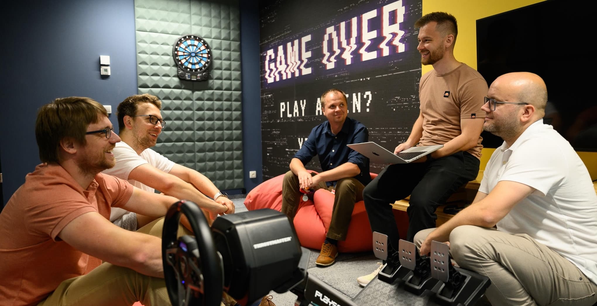 Five colleagues in a casual meeting room with a “Game Over” wall sign, chatting on bean bags and one with a laptop.