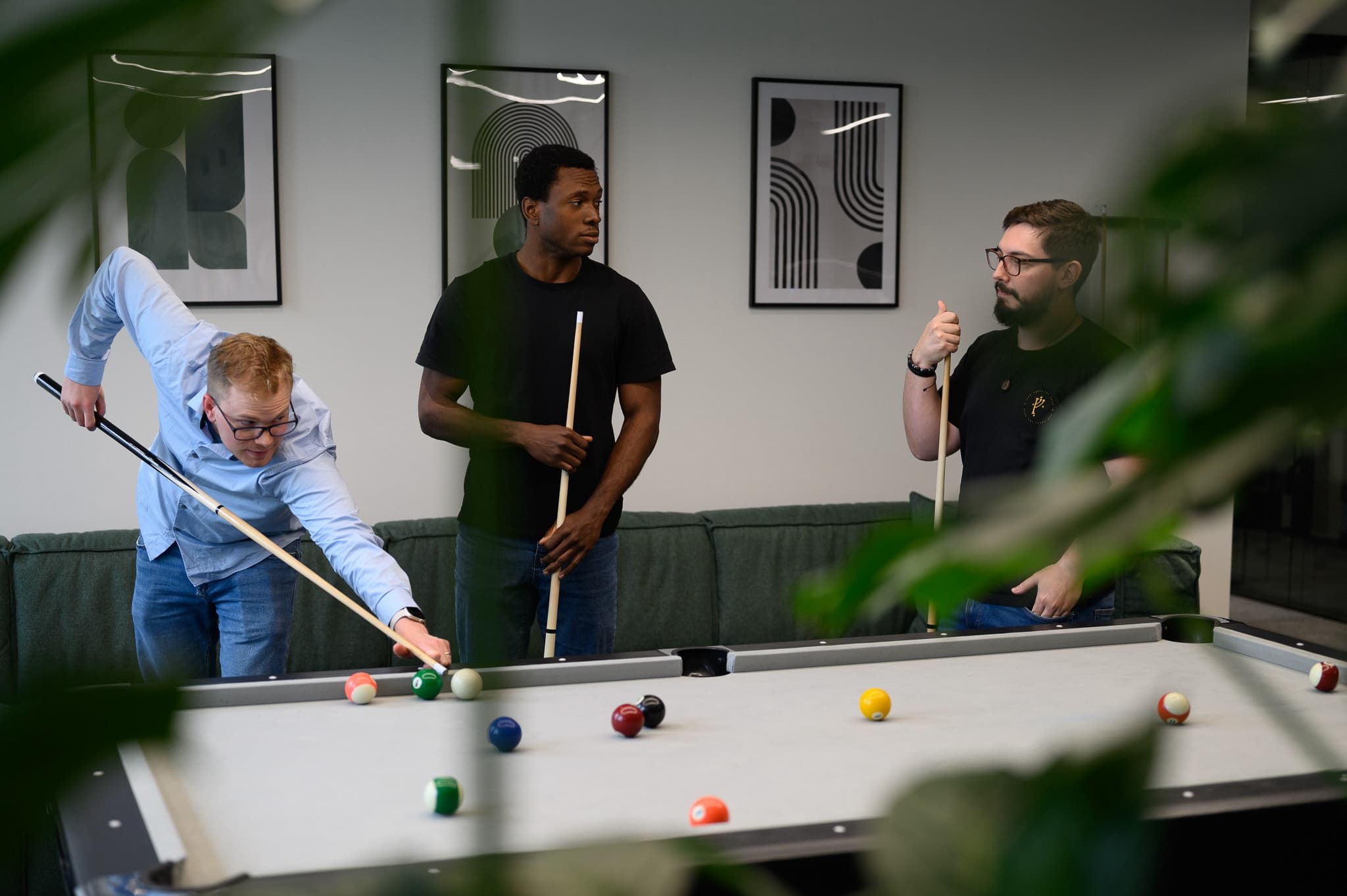 Three men playing pool in an office break area, one leaning to take a shot while the others watch with cues in hand.