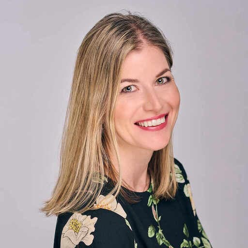 Woman smiling with blonde hair wearing a dark floral blouse, posed against a plain light grey background.