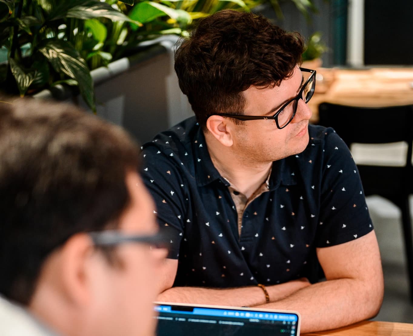 Man in a navy patterned shirt with glasses looks to the right, seated at a wooden table with greenery in the background.
