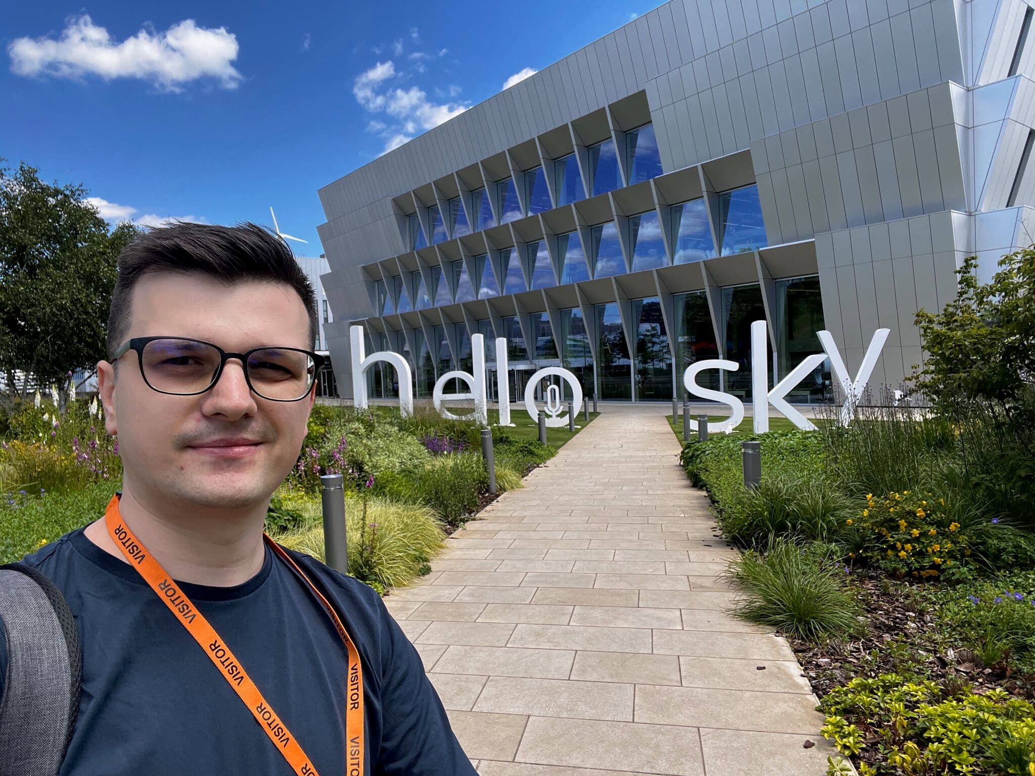 Man with glasses taking a selfie outside a modern building with “hello sky” signage in the garden path.