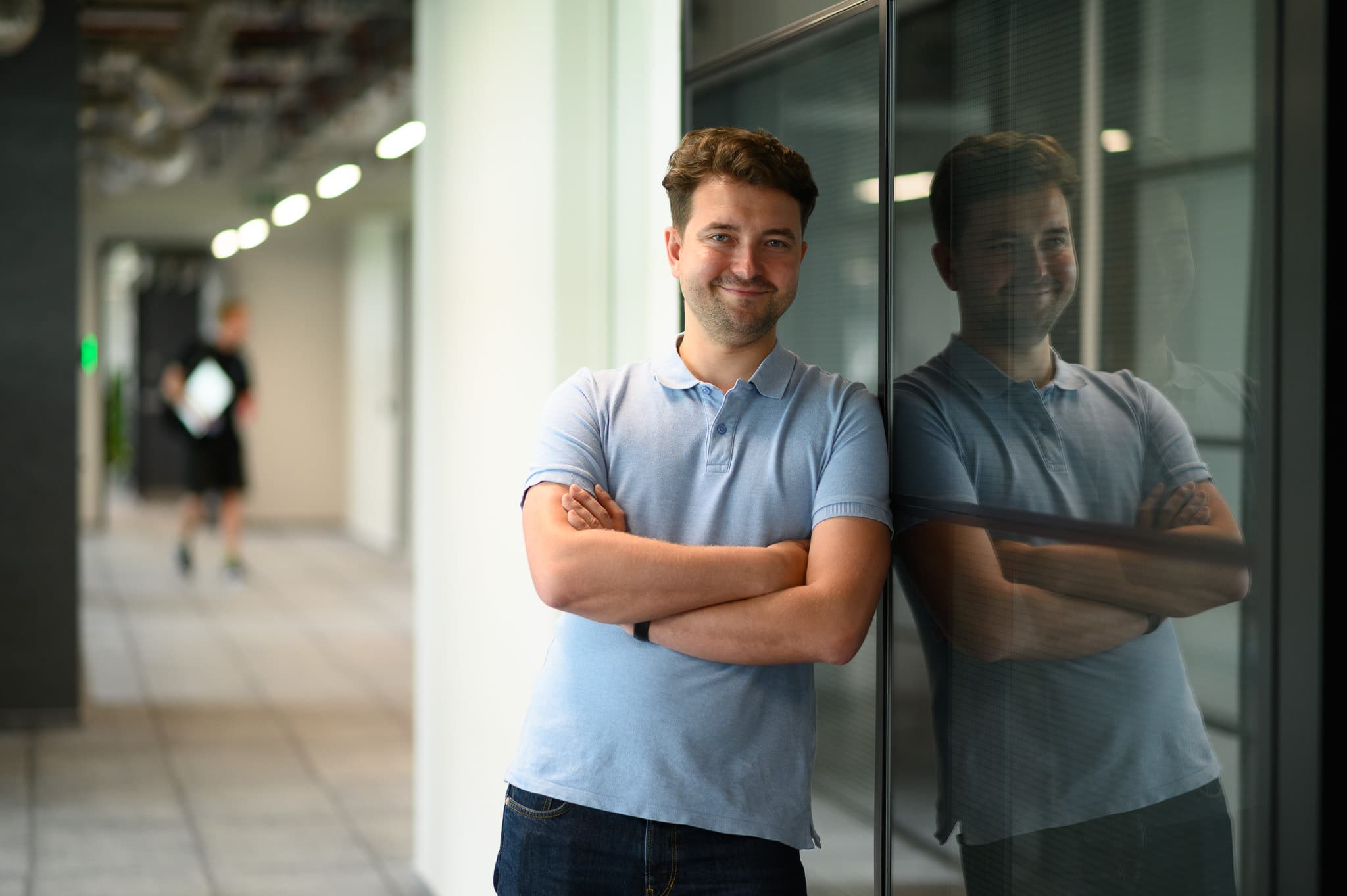 Man standing with arms crossed and smiling, leaning against a glass wall with his reflection visible.