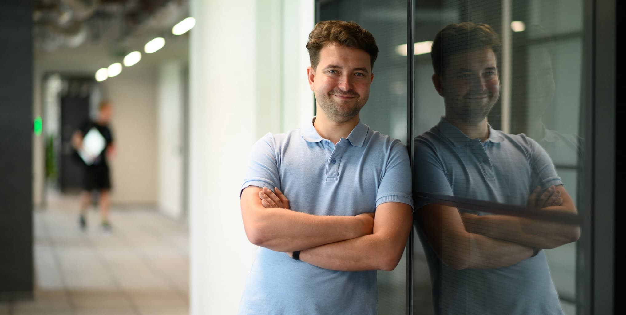 Man standing with arms crossed and smiling, leaning against a glass wall with his reflection visible.
