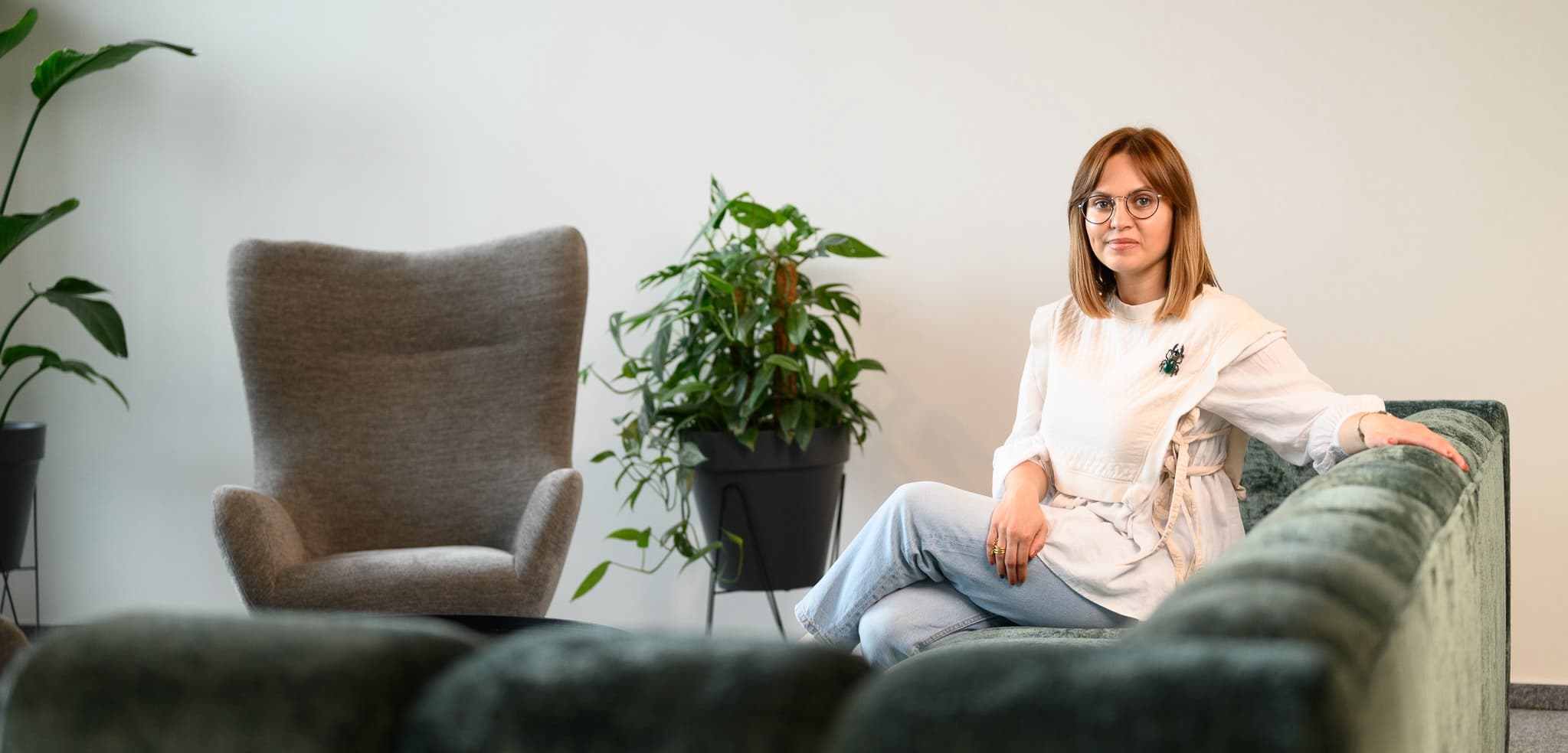 Woman seated on a velvet sofa, posing confidently for a photo with houseplants and armchairs in the background.