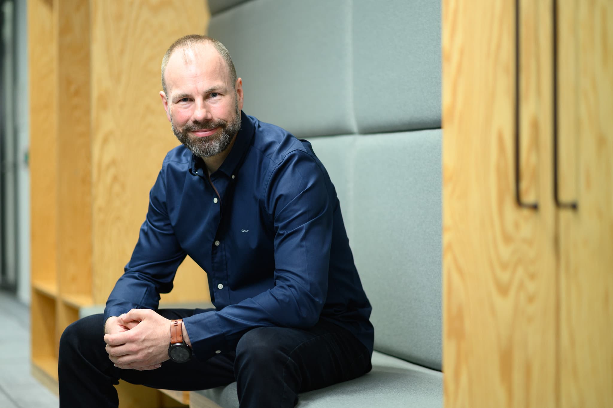 Bearded man in a dark blue shirt seated on a bench in a modern office hallway, smiling at the camera.
